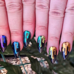 Può includere: Primo piano di una mano con unghie a stiletto nere con un effetto gocciolante. Le unghie sono dipinte di nero con un design a goccia dorato, verde e blu metallizzato.
