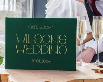 May include: A green wedding guest book with gold lettering that reads "Kate & John Wilsons Wedding 10.01.2024". The book is sitting on a wooden table with two champagne flutes.