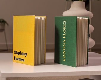 Op de afbeelding: Twee hardcover boeken op een witte tafel. Eén boek is geel met de naam "Stephany Fuentes" in zwart. Het andere boek is groen met "KRISTINA FLORES" in goud. Beide boeken zijn open en tonen witte pagina's.