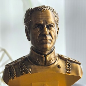 May include: A gold-colored bust sculpture of a man wearing a military uniform. The bust is on a yellow base with a decorative design.