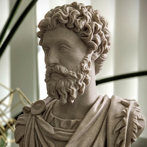 May include: A detailed bust of a man with curly hair and a beard, sculpted in a light beige color. The sculpture features draped fabric over the shoulders and chest, with intricate detailing. The bust is placed on a dark surface.