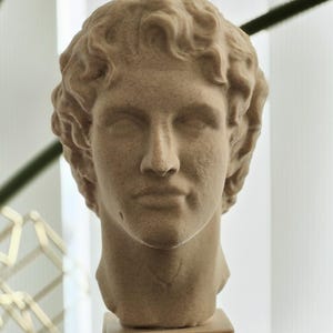 May include: A beige-colored bust of a man with curly hair, set on a square pedestal. The sculpture has a classical style, with detailed facial features and a smooth texture. The bust is displayed on a dark surface.