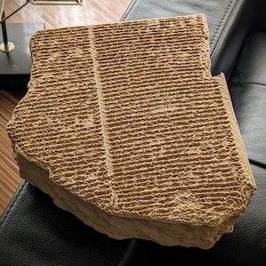 May include: A tan, rectangular stone-like tablet featuring a textured surface with numerous horizontal, parallel grooves and a single vertical line running down the centre, mimicking the appearance of an ancient cuneiform clay artefact.