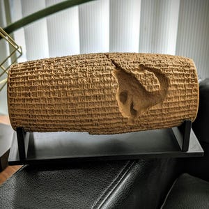 Puede incluir: Un artefacto cilíndrico de color beige con intrincadas tallas y una figura de animal estilizada. El objeto se exhibe sobre un soporte negro, lo que sugiere un contexto museístico o histórico. La textura y el color sugieren que está hecho de arcilla o piedra.