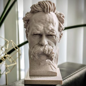 May include: A light beige stone bust of Friedrich Nietzsche. The sculpture shows a detailed depiction of Nietzsche's face, with a prominent mustache and furrowed brow. The bust is mounted on a rectangular base with the name "Nietzsche" inscribed.