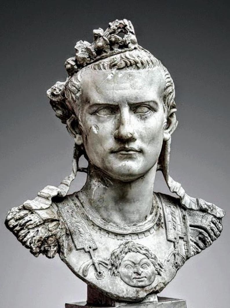 K&ouml;nnte beinhalten: Eine wei&szlig;e Marmorb&uuml;ste einer Person mit einem Bl&auml;tterkranz und einer R&uuml;stung. Der Brustpanzer zeigt ein zentrales Relief eines Gesichts. Die Figur hat einen ernsten Ausdruck, markante Gesichtsz&uuml;ge und B&auml;nder, die seitlich am Kopf herabh&auml;ngen.