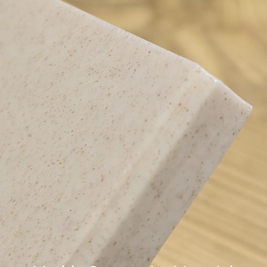 K&ouml;nnte beinhalten: Ein Eckabschnitt einer Platte aus cremefarbenem Marmor-Verbundwerkstoff mit feinen braunen Sprenkeln auf der gesamten Oberfl&auml;che. Das Material hat eine glatte, matte Oberfl&auml;che und eine saubere, gerade Kante, mit dem Text Marble Composite Material.