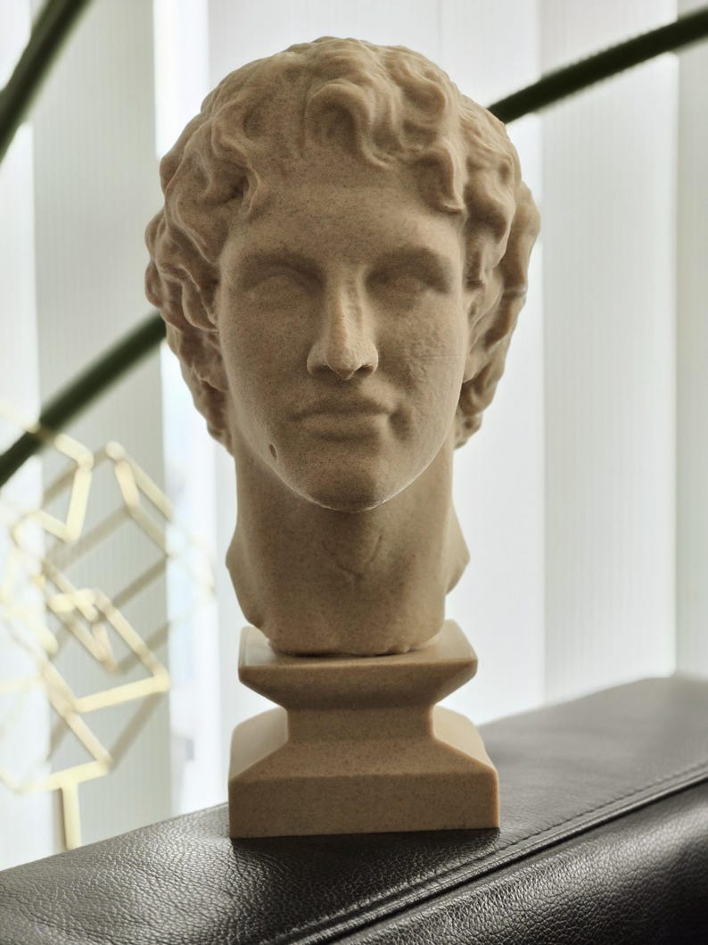 May include: A beige-colored bust of a man with curly hair, set on a square pedestal. The sculpture has a classical style, with detailed facial features and a smooth texture. The bust is displayed on a dark surface.