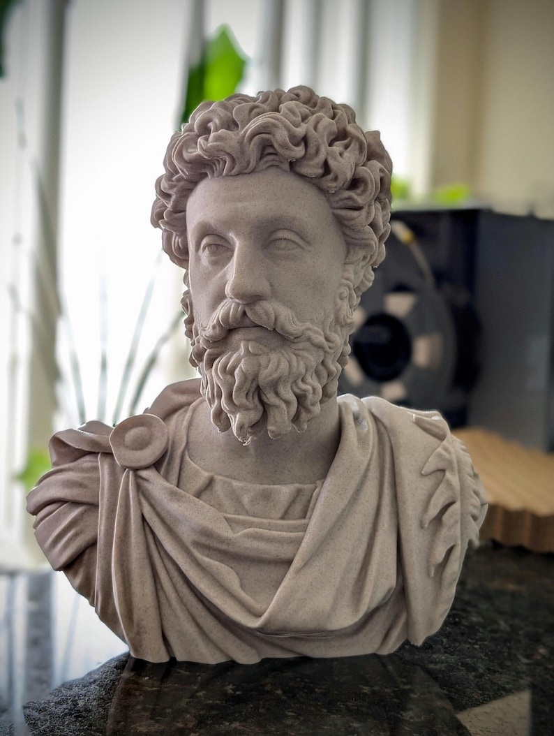 May include: A detailed bust of a man with curly hair and a beard, sculpted in a light beige color. The sculpture features a draped garment and is likely a classical or historical figure.