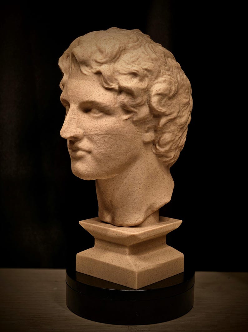 May include: A tan-colored bust of a classical figure, featuring detailed facial features and curly hair. The sculpture is mounted on a square pedestal and a black circular base, set against a dark background. The bust is a decorative item.