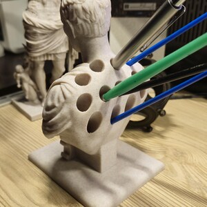 May include: A light beige bust of a classical figure with multiple holes, used as a pen holder. Several pens in various colours, including silver, green, blue, and black, are inserted into the holes. The bust is on a square base.