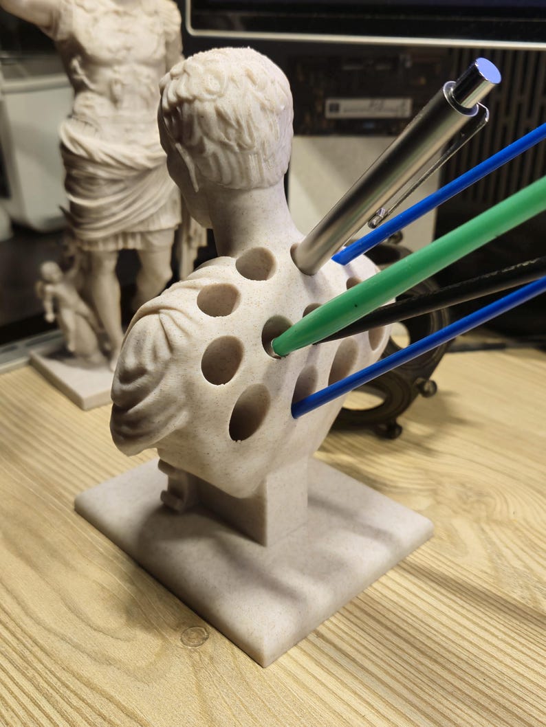 May include: A light beige bust of a classical figure with multiple holes, used as a pen holder. Several pens in various colours, including silver, green, blue, and black, are inserted into the holes. The bust is on a square base.