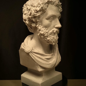 May include: A detailed bust of a man with curly hair and a beard, sculpted in a light beige color. The sculpture features a draped garment and is mounted on a black base, set against a dark background.
