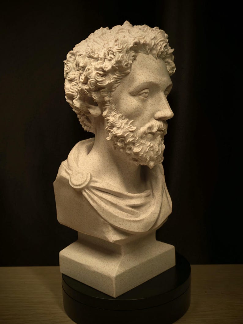 May include: A detailed bust of a man with curly hair and a beard, sculpted in a light beige color. The sculpture features a draped garment and is mounted on a black base, set against a dark background.