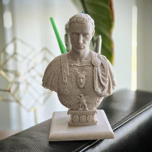 May include: A beige Roman emperor bust, with intricate details of the face and clothing. The sculpture sits on a square base with floral patterns. This decorative item is likely for home decor, and is measured in centimeters.