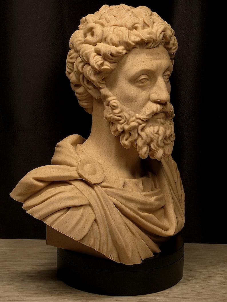 May include: A tan-colored bust of a man with a curly hairstyle and a full beard. The sculpture depicts a Roman emperor, wearing a draped garment fastened with a circular clasp. The bust is mounted on a black base, showcasing classical art.
