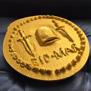 May include: A gold-coloured round plaque with a raised design of a helmet, two swords, and the text "Eid-Mar".