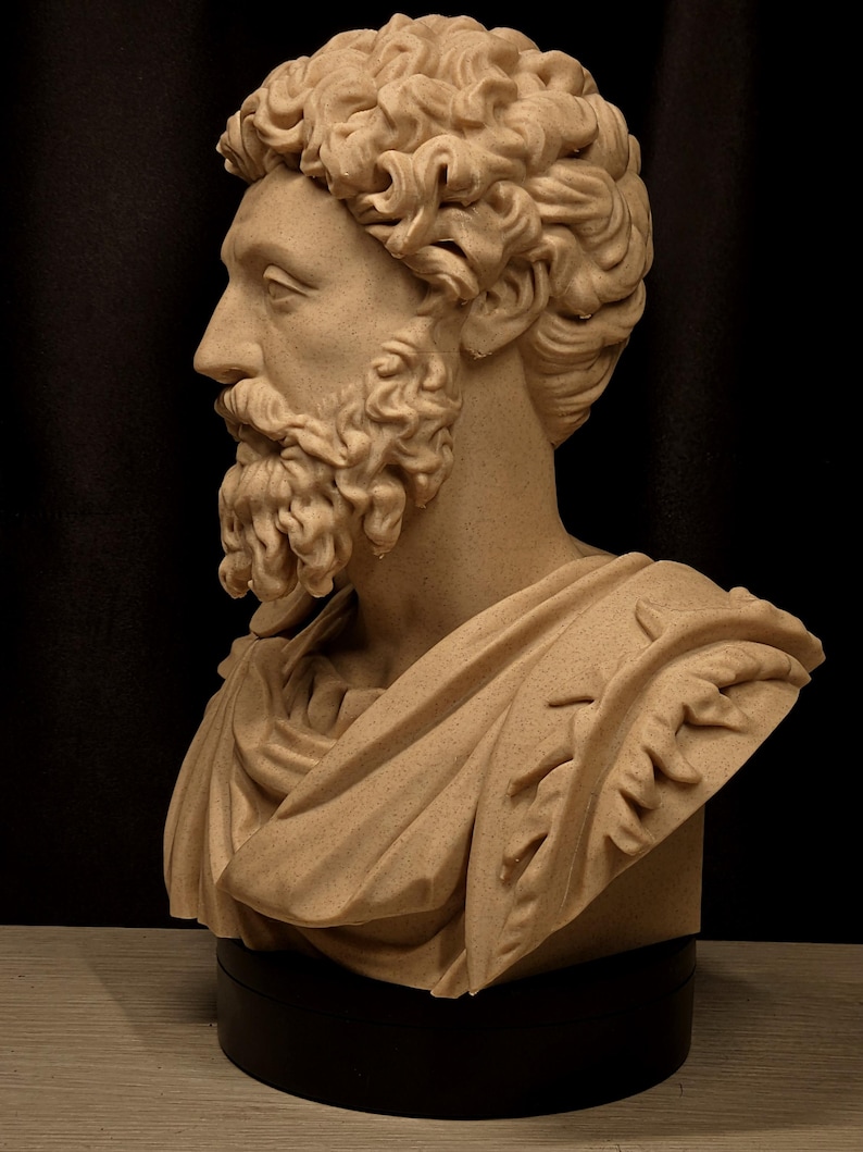 May include: A tan-colored bust of a man with curly hair and a beard, sculpted in the classical style. The bust features detailed facial features and is draped with a toga. The sculpture is mounted on a black base, set against a dark background.
