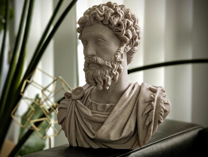 May include: A detailed bust of a man with curly hair and a beard, sculpted in a light beige color. The sculpture features draped fabric over the shoulders and chest, with intricate detailing. The bust is placed on a dark surface.