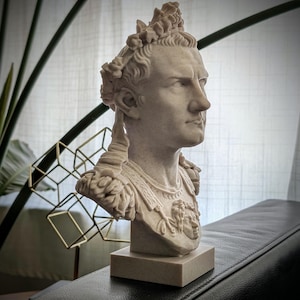 K&ouml;nnte beinhalten: Eine hellbeige B&uuml;ste eines r&ouml;mischen Kaisers mit Lorbeerkranz und verzierter R&uuml;stung, die auf der Brust ein Gorgoneion-Emblem zeigt. Die Figur ist im Profil dargestellt und steht auf einem quadratischen Sockel mit detaillierten Z&uuml;gen.