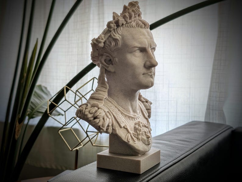 K&ouml;nnte beinhalten: Eine hellbeige B&uuml;ste eines r&ouml;mischen Kaisers mit Lorbeerkranz und verzierter R&uuml;stung, die auf der Brust ein Gorgoneion-Emblem zeigt. Die Figur ist im Profil dargestellt und steht auf einem quadratischen Sockel mit detaillierten Z&uuml;gen.