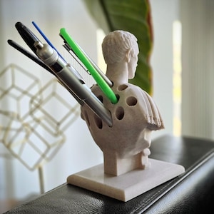 May include: A beige pen holder in the shape of a bust, with multiple holes for pens. Various pens, including black, blue, and green, are inserted. The sculpture sits on a square base. The background is out of focus.