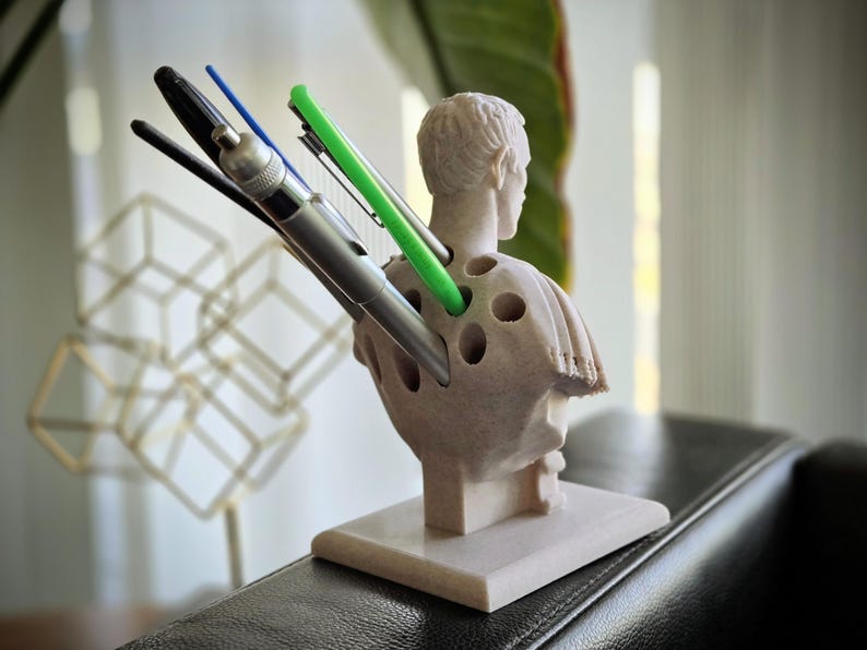 May include: A beige pen holder in the shape of a bust, with multiple holes for pens. Various pens, including black, blue, and green, are inserted. The sculpture sits on a square base. The background is out of focus.