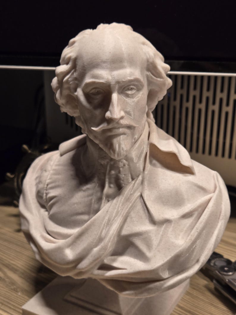 May include: A white 3D printed bust of William Shakespeare. The bust is detailed and shows Shakespeare's face and shoulders. The bust is on a white base.