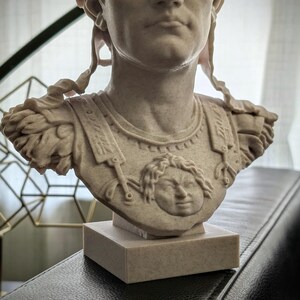 K&ouml;nnte beinhalten: Eine beige, steinstrukturierte B&uuml;ste einer r&ouml;mischen Figur in R&uuml;stung mit einem zentralen Gorgonenhaupt-Relief. Die Skulptur zeigt detaillierte Schulterst&uuml;cke und einen rechteckigen Sockel, die den Oberk&ouml;rper und Kopf darstellen.