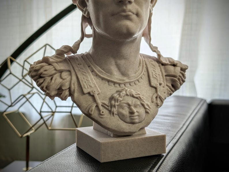K&ouml;nnte beinhalten: Eine beige, steinstrukturierte B&uuml;ste einer r&ouml;mischen Figur in R&uuml;stung mit einem zentralen Gorgonenhaupt-Relief. Die Skulptur zeigt detaillierte Schulterst&uuml;cke und einen rechteckigen Sockel, die den Oberk&ouml;rper und Kopf darstellen.
