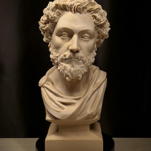 May include: A beige-colored bust sculpture of a man with curly hair and a beard. The sculpture features a draped garment over the shoulders and is mounted on a square base. The bust is set against a dark background.