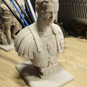 May include: A light beige bust of a Roman emperor, with detailed armour and a square base. The sculpture has intricate detailing on the face, hair, and clothing, showcasing a classical art style. The bust is a decorative item.
