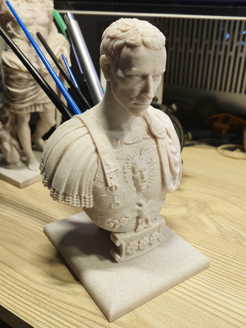 May include: A light beige bust of a Roman emperor, with detailed armour and a square base. The sculpture has intricate detailing on the face, hair, and clothing, showcasing a classical art style. The bust is a decorative item.