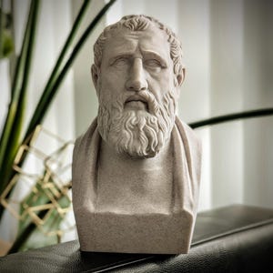 May include: A stone-coloured bust of a bearded man with a serious expression. The sculpture features detailed facial features, including a prominent nose and a full beard. The bust is set against a blurred background with greenery.
