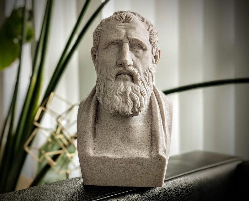 May include: A stone-coloured bust of a bearded man with a serious expression. The sculpture features detailed facial features, including a prominent nose and a full beard. The bust is set against a blurred background with greenery.
