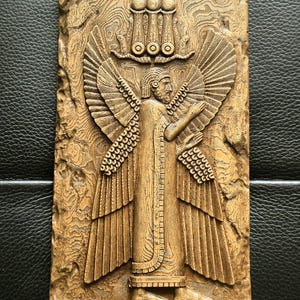 May include: A detailed wooden relief carving of a winged figure with outstretched arms, wearing a long robe and a headdress. The figure has large, detailed wings and stands against a textured background. The relief is in shades of brown.