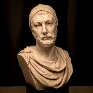 May include: A stone bust of a man with a beard and a detailed helmet. The sculpture is set against a dark backdrop. The man has a serious expression and is wearing a draped garment. The bust is a light beige colour.