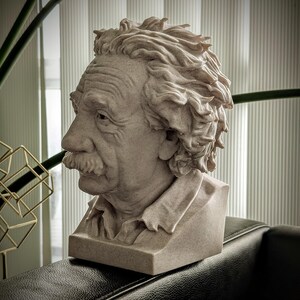 May include: A beige bust sculpture of Albert Einstein, featuring detailed facial features including a mustache and textured hair. The sculpture is set on a rectangular base and is displayed on a dark surface.