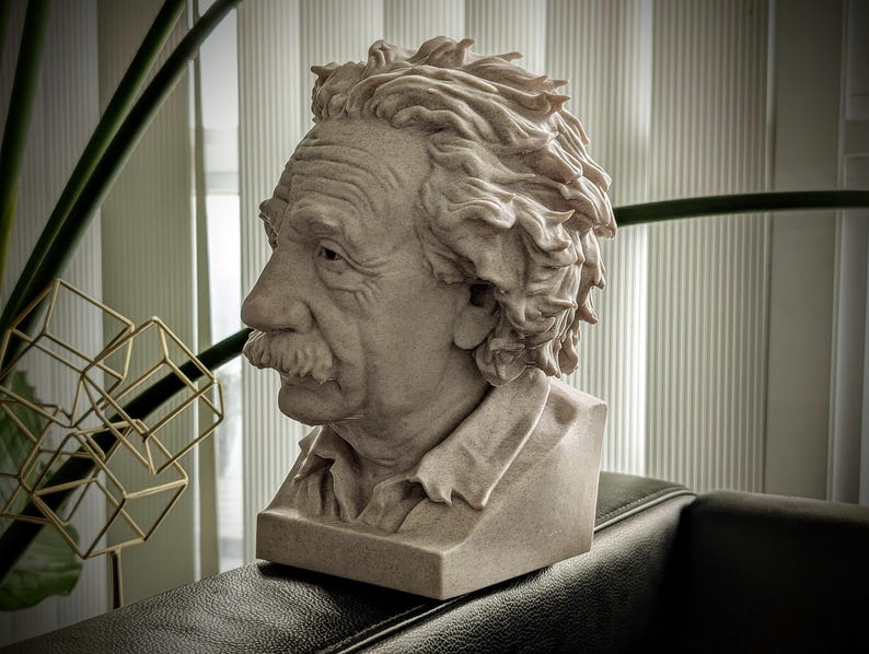 May include: A beige bust sculpture of Albert Einstein, featuring detailed facial features including a mustache and textured hair. The sculpture is set on a rectangular base and is displayed on a dark surface.