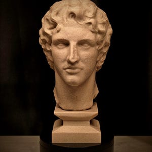 May include: A beige-colored bust of a classical male figure with curly hair. The sculpture is mounted on a square pedestal and a black circular base, set against a dark background. The bust has detailed facial features.