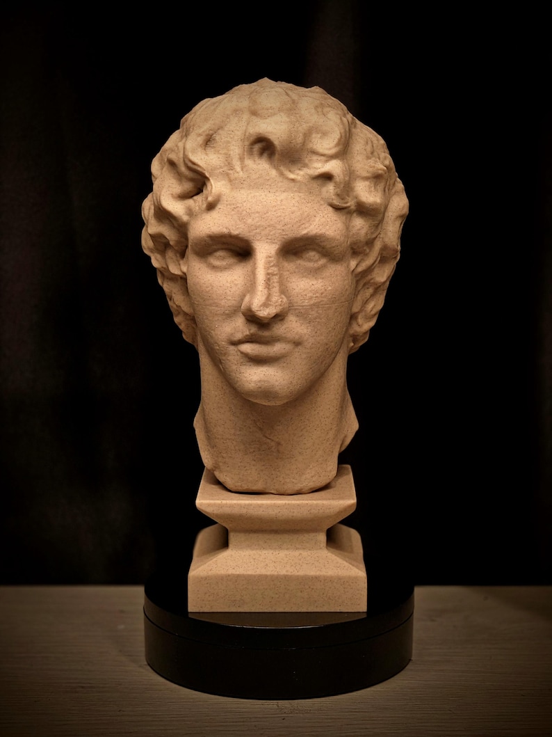 May include: A beige-colored bust of a classical male figure with curly hair. The sculpture is mounted on a square pedestal and a black circular base, set against a dark background. The bust has detailed facial features.