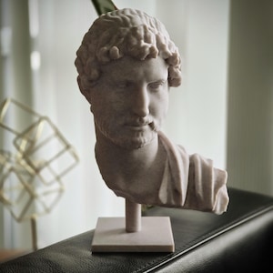 Puede incluir: Busto de color claro de un emperador romano, con rasgos faciales detallados y cabello rizado. La escultura est&aacute; montada sobre una base cuadrada. El busto es un objeto decorativo. Est&aacute; sobre una superficie oscura.
