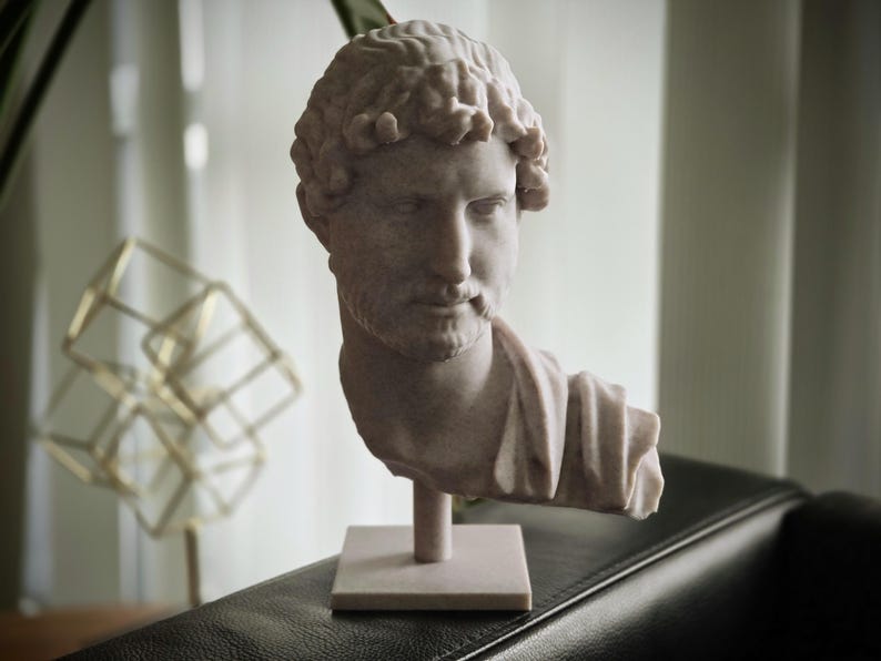 Puede incluir: Busto de color claro de un emperador romano, con rasgos faciales detallados y cabello rizado. La escultura est&aacute; montada sobre una base cuadrada. El busto es un objeto decorativo. Est&aacute; sobre una superficie oscura.