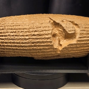 May include: A tan, cylindrical clay-like artifact featuring dense, horizontal rows of cuneiform script carved into its surface. A prominent, irregular indentation or chip is visible on the right side of the cylinder, which rests on a black stand.
