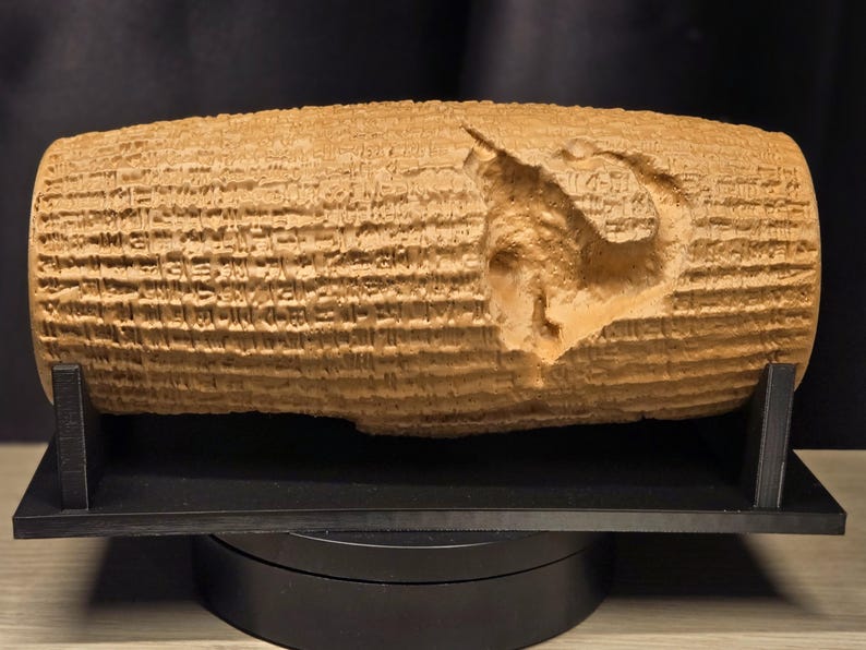 May include: A tan, cylindrical clay-like artifact featuring dense, horizontal rows of cuneiform script carved into its surface. A prominent, irregular indentation or chip is visible on the right side of the cylinder, which rests on a black stand.