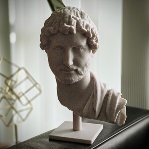 Puede incluir: Busto de color claro de un hombre con cabello rizado y barba, montado sobre una base cuadrada. La escultura es de estilo cl&aacute;sico, posiblemente romano o griego, y est&aacute; hecha de un material similar a la piedra. El busto est&aacute; sobre un fondo borroso.