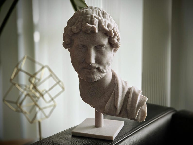 Puede incluir: Busto de color claro de un hombre con cabello rizado y barba, montado sobre una base cuadrada. La escultura es de estilo cl&aacute;sico, posiblemente romano o griego, y est&aacute; hecha de un material similar a la piedra. El busto est&aacute; sobre un fondo borroso.