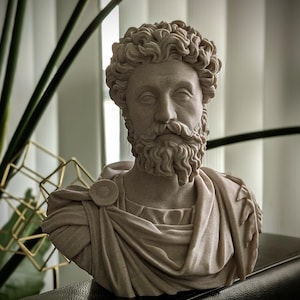 May include: A detailed bust of a man with curly hair and a beard, sculpted in a light beige color. The sculpture depicts a classical figure, wearing draped clothing and a circular brooch. The bust is a decorative item.