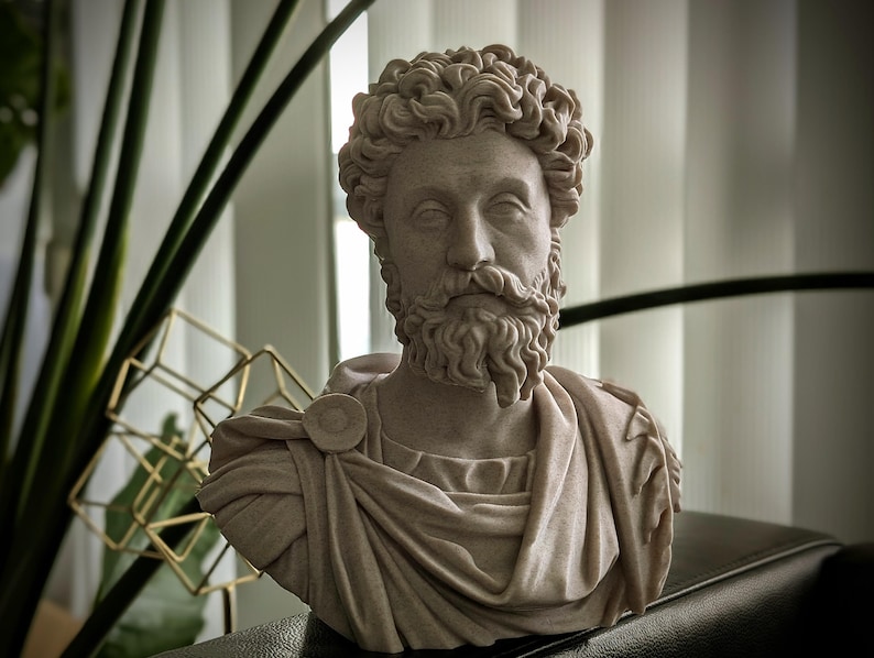 May include: A detailed bust of a man with curly hair and a beard, sculpted in a light beige color. The sculpture depicts a classical figure, wearing draped clothing and a circular brooch. The bust is a decorative item.