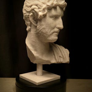 Puede incluir: Busto beige de un hombre con cabello rizado y barba, montado sobre una base cuadrada y un soporte cil&iacute;ndrico. La escultura se encuentra sobre un fondo negro, resaltando los detalles cl&aacute;sicos.